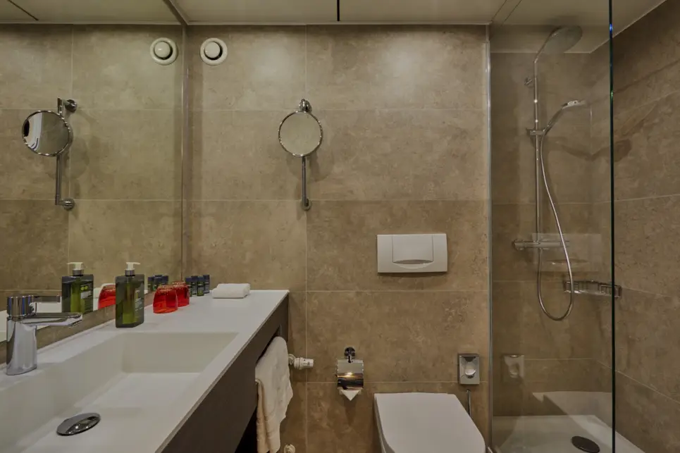 Badezimmer mit einem großem Waschbecken und einem Spiegel auf der linken Seite. AUf der rechten Seite ist eine Dusche.