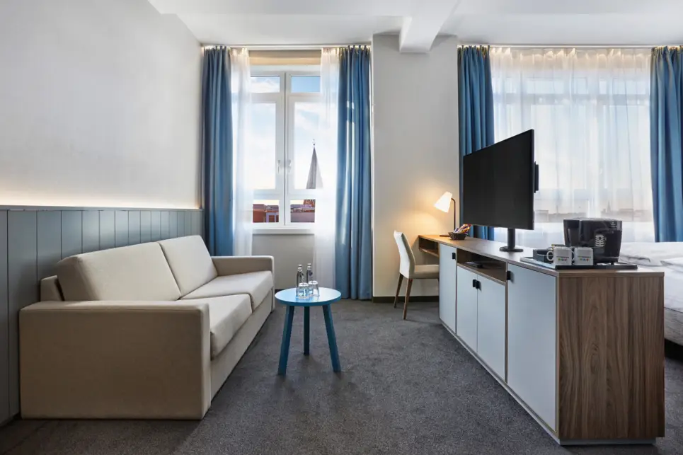 Hotelzimmer mit einer Couch auf der linken Seite und einer kleinen Kommode mit einem Fernseher auf der rechten Seite. Vor der Couch steht ein kleiner Tisch.