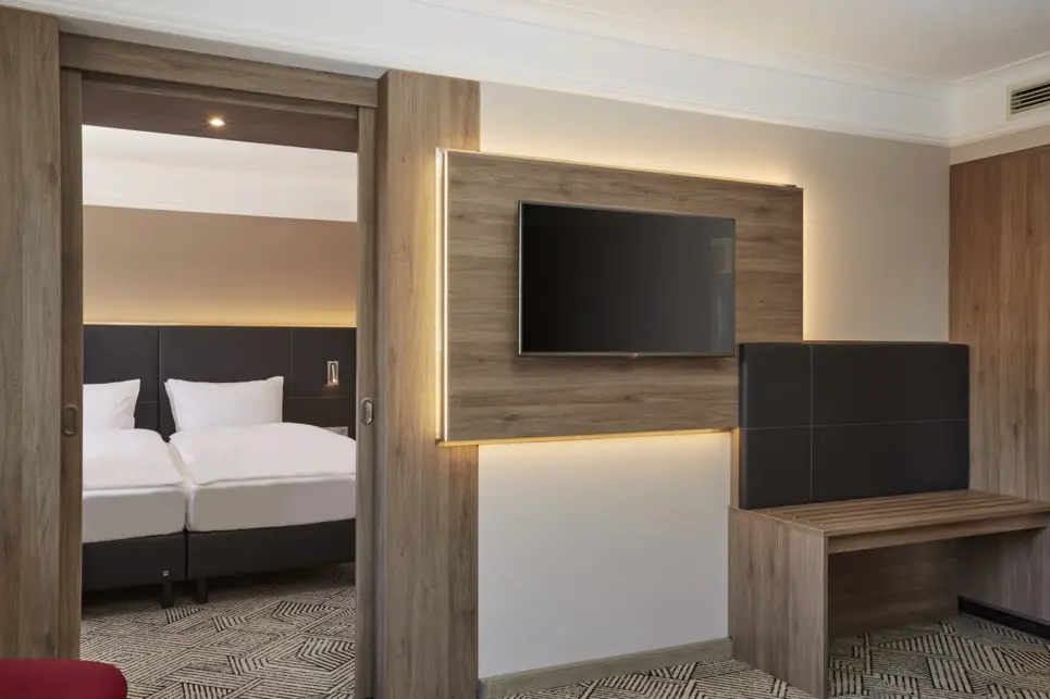 Hotelzimmer mit einem Flachbildfernseher und einer Taschenablage an der Wand. Durch eine Tür sieht man ein Doppelbett in einem anderen Raum.
