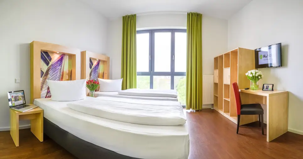 Zwei Einzelbetten vor einem großen Fenster. Vor den Betten steht ein Schrank und ein Schreibtisch mit einem Stuhl.