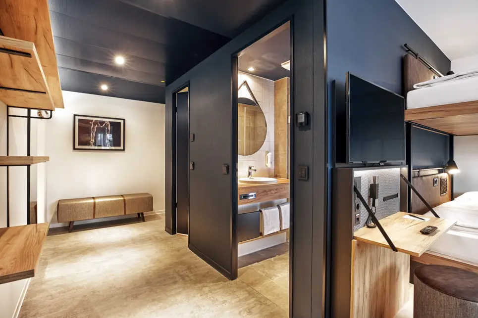 Blick vom Zimmer in den Zimmerflur mit einer Bank und einem Regal. Die Tür zum Badezimmer ist offen und man sieht ein Waschbecken.