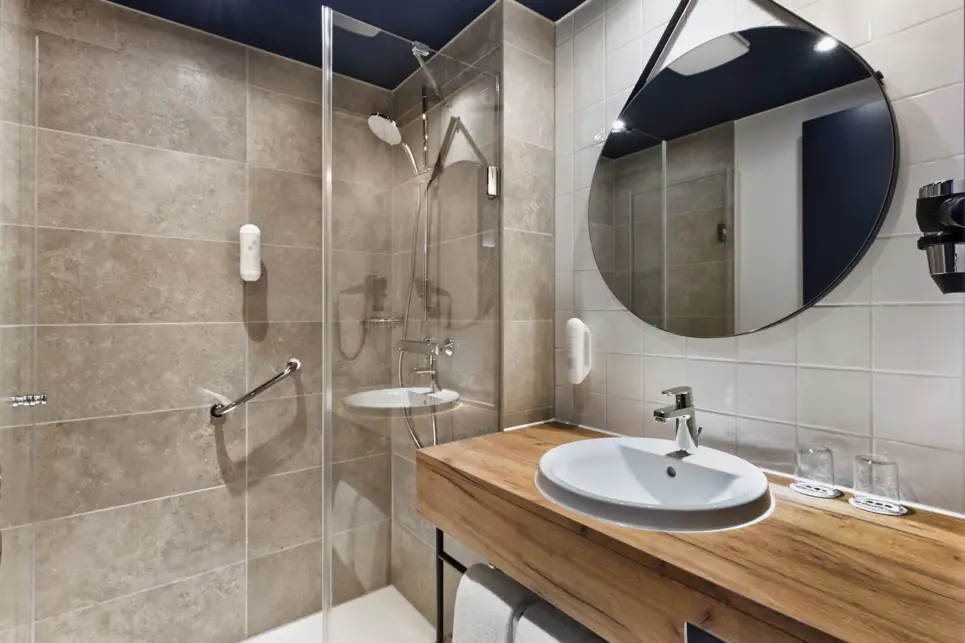 Badezimmer mit einer großen Dusche. Neben der Dusche ist ein brauner Waschtisch mit Waschbecken und einem großen Spiegel darüber.