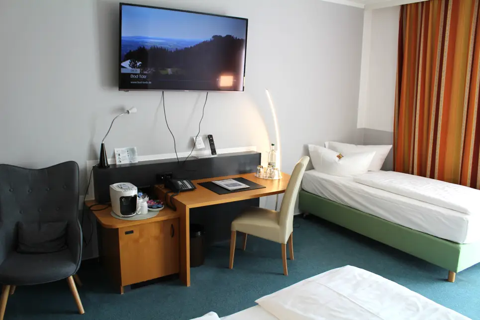Hotelzimmer mit einem Schreibtisch und einem Fernseher an der Wand. Neben dem Schreibtisch steht auf der rechten Seite ein Einzelbett.