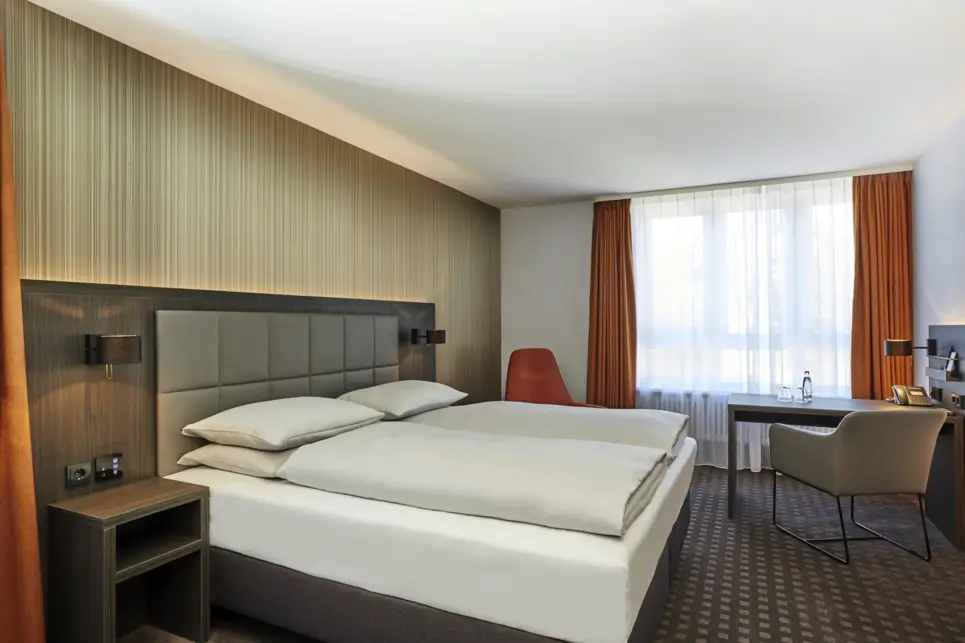 Auf der linken Seite sthet ein Doppelbett. Auf der rechten Seite steht ein kleiner Schreibtisch mit einem Stuhl.