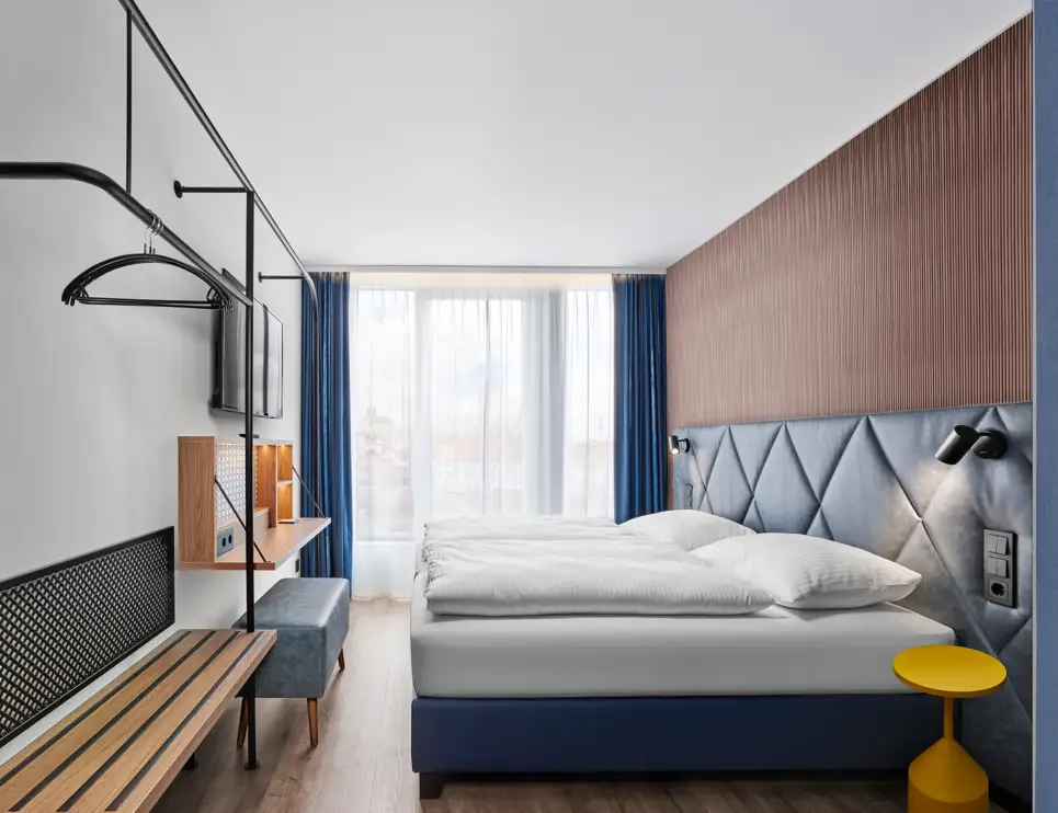 Hotelzimmer mit einem Doppelbett vor einem großen Fenster mit Gardine und offenen Vorhängen. Links vor dem Bett hängt ein Fernseher an der Wand, darunter befindet sich ein aufgeklappter Tisch und ein breiter Hocker. 