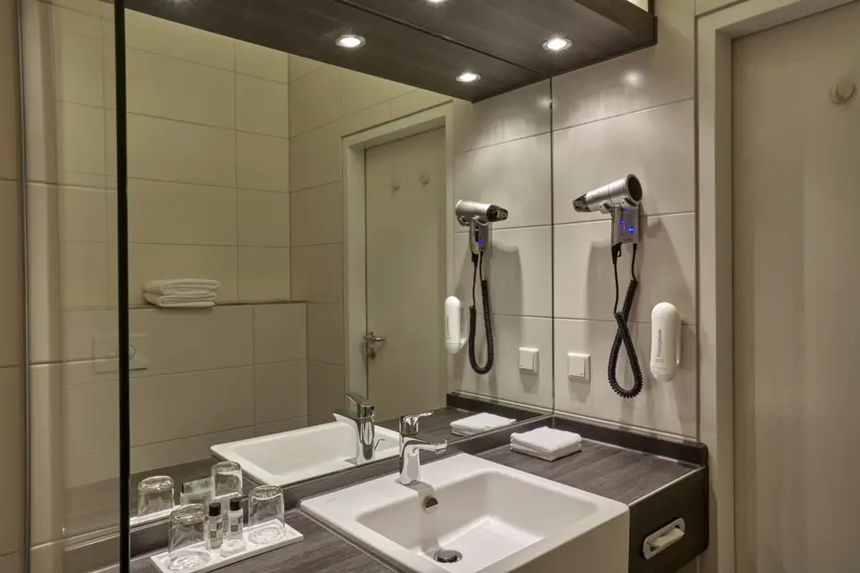 Waschbecken vor einem großen Spiegel im Badezimmer. Daneben hängt ein Föhn.