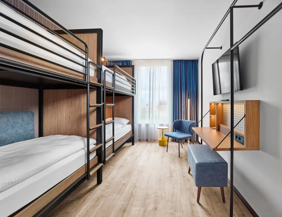 Etagenbetten im Hotelzimmer. In der Ecke steht ein Stuhl mit einem Tisch. Rechts an der Wand hängt ein Fernseher, darunter ist ein aufgeklappter Schreibtisch und ein breiter Sitzhocker.