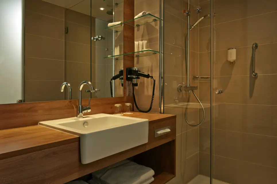 Badezimmer mit großem Waschtisch und großem Spiegel auf der linken Seite. Auf der rechten Seite dahinter ist eine große, begehbare Dusche mit Glaswand.