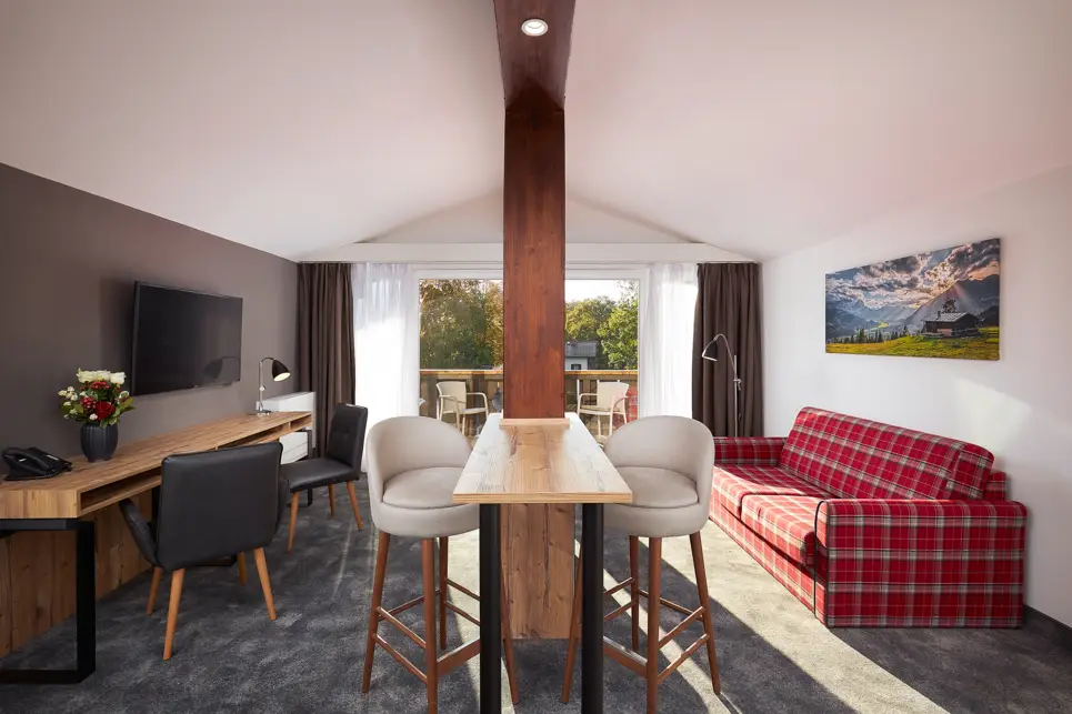 Blick aus dem Zimmer auf den Balkon und das Bergpanorama. Auf der linken Seite steht ein Schreibtisch mit 2 Stühlen vor einem hängenden TV an der Wand. Rechts steht ein weiß-rot kariertes Sofa an der Wand.