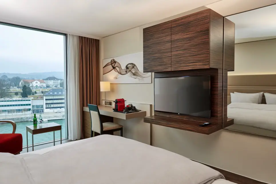 Hotelzimmer mit Doppelbett und großem Fenster mit Blick auf die Aaare. Am Fußende  hängt ein Fernseher und ein großer Spiegel an der Wand.