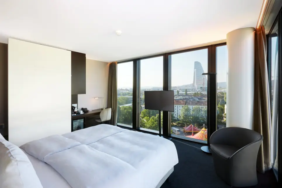 Blick vom Doppelbett in das Zimmer mit einem Schreibtisch auf der linken Seite. Am Fußende steht ein Fernseher und dahinter bodentiefe Fenster.