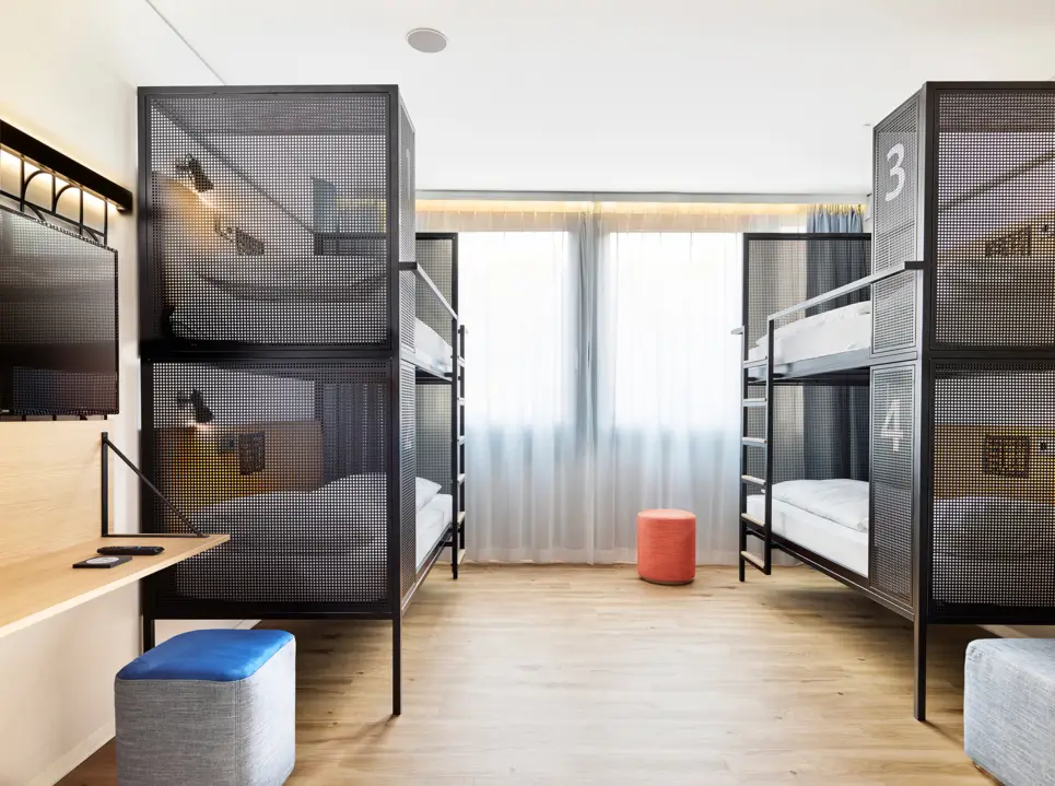 Blick in das Hotelzimmer mit zwei Etagenbetten. Vor den Betten stehen Hocker, ein Schreibtisch und ein Fernseher.