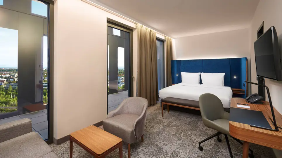 Im Hintergrund steht ein Doppelbett. Im Vordergrund auf der rechten Seite ein Schreibtisch mit Stuhl und auf der linken Seite bodentiefe Fenster.