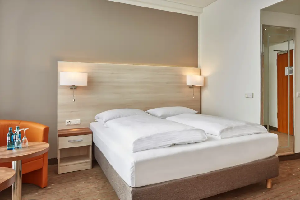 Hotelzimmer mit Doppelbett, Sessel und einem Tisch mit Wasserflaschen und Gläsern.