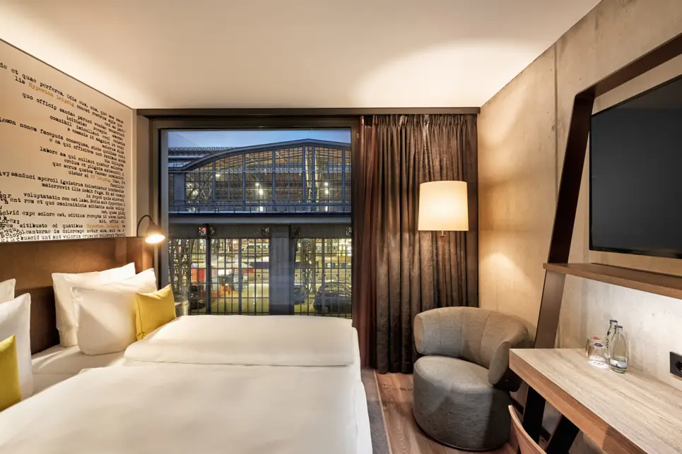 Hotelzimmer bei Nacht mit Blick in den Hauptbahnhof. Auf der linekn Seite steht ein Doppelbett, auf der rechten Seite ein Tisch mit einem Fernseher.