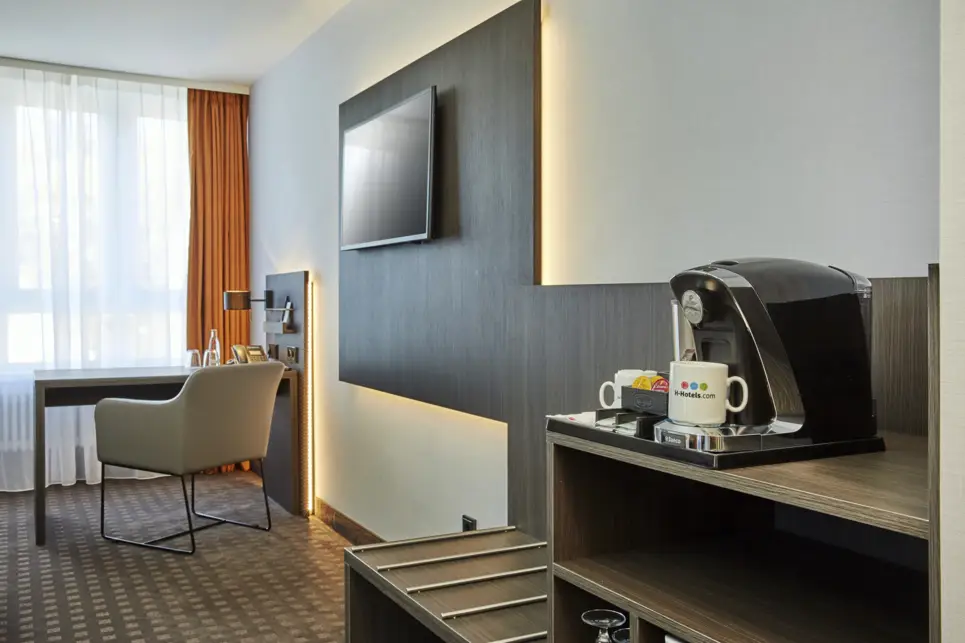 Hotelzimmer mit einem Schreibtisch auf der linken Seite, in der Mitte hängt ein Fernseher und auf der rechten Seite eine Kaffeemaschine.