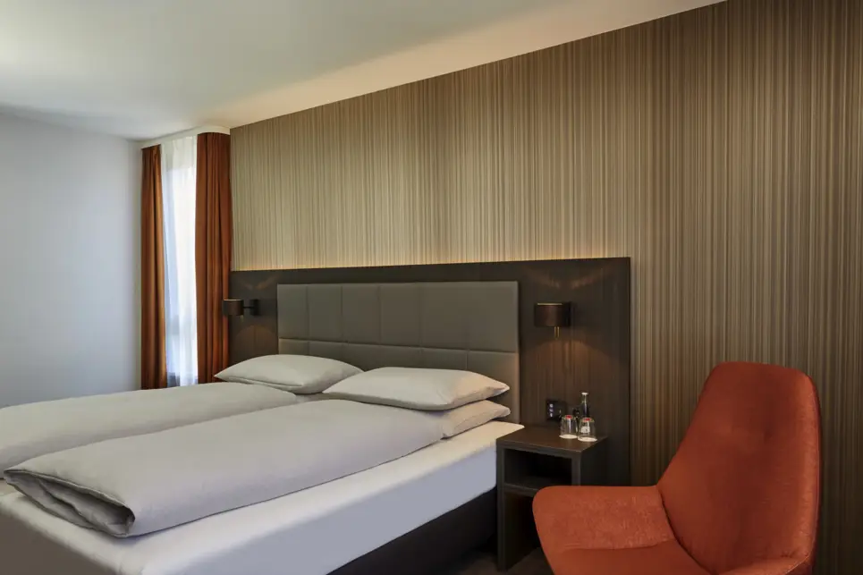 Doppelbett vor einer goldgestreiften Wand. Daneben steht ein roter Stuhl.