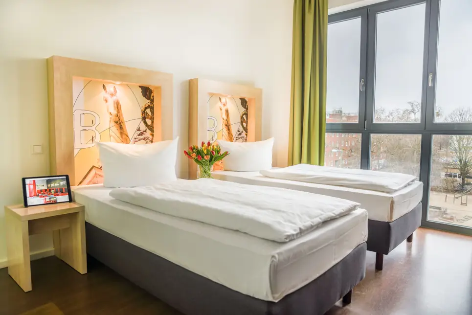 Zwei Einzelbetten vor einem großen Fenster. Neben den Betten steht ein kleiner Nachttisch.