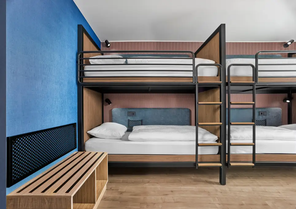 Blick auf Etagenbetten mit Leitern. Links vor den Betten ist ein Holzgestell zum Sitzen oder zum Abstellen von Taschen.