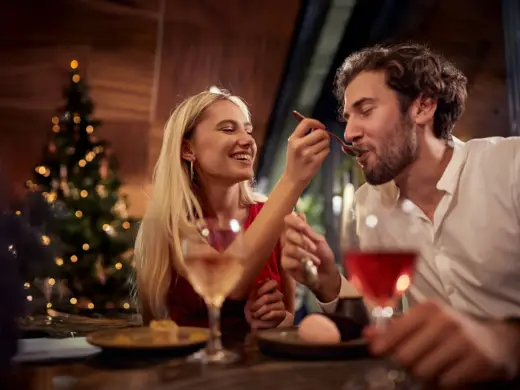 Eine Frau füttert einen Mann mit einem goldenen Löffel. Im Hintergrund steht ein Weihnachtsbaum.