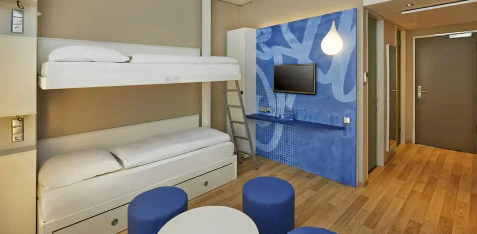 Hotelzimmer mit einem Etagenbett und einer Sitzgruppe. Vor dem Bett hängt ein Fernseher und ein kleiner Schreibtisch.