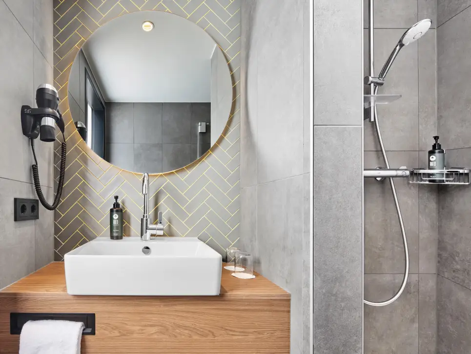 Eckiges Waschbecken auf Holztisch mit rundem Wandspiegel und Föhn. Rechts daneben ist eine Dusche.