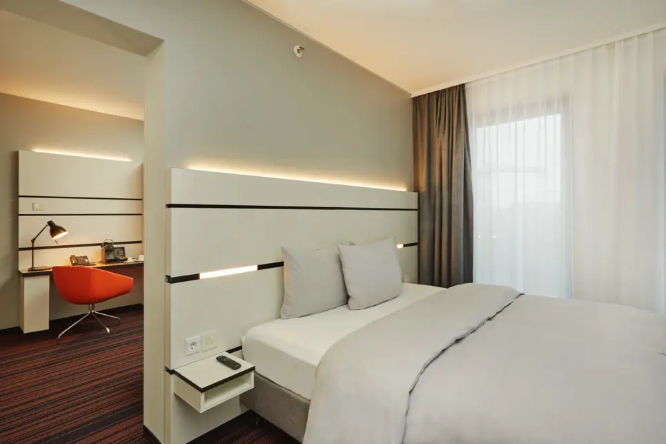 Schlichtes, modernes Zimmer in Schwarzweiß und beleuchtetem Kopfteil. Hinter der Wand am Bett ist ein zweites Zimmer mit Schreibtisch und einem roten Stuhl.