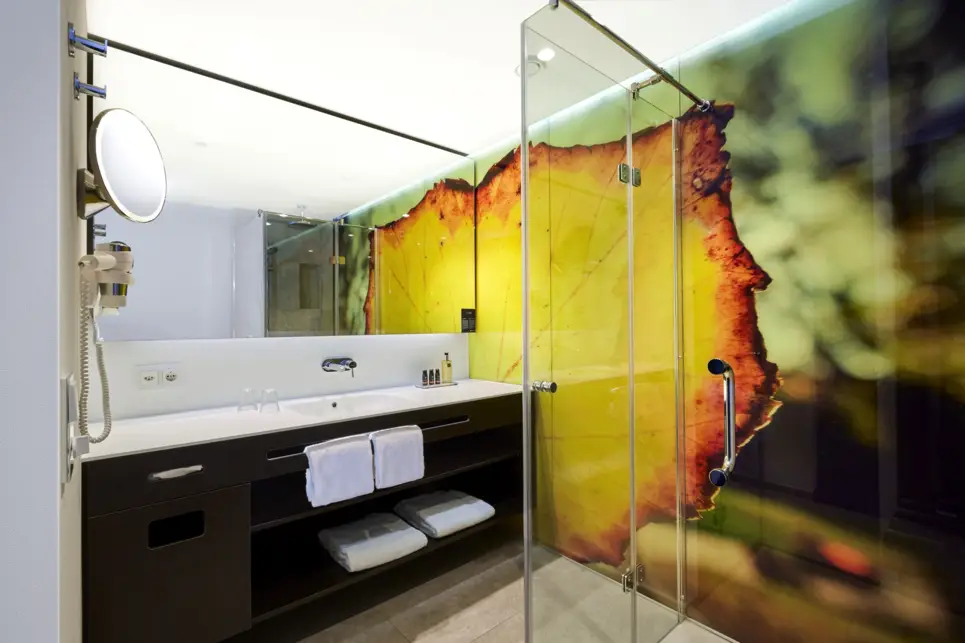 Badezimmer mit einer grünen Tapete mit einem Blatt. Auf der rechten Seite steht eine Dusche und auf der linken Seite ein Waschbecken mit einem großen Spiegel.