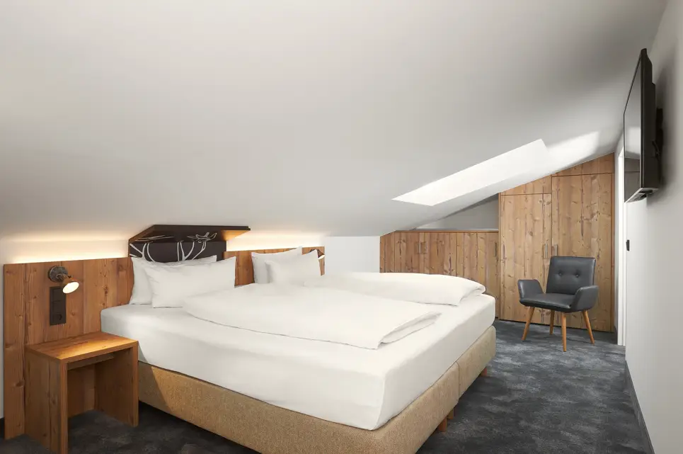 Hotelzimmer mit einem großen Doppelbett auf der linken Seite und einem Fernseher auf der rechten Seite an der Wand. Im Hintergrund steht ein Stuhl vor einem großen Schrank.