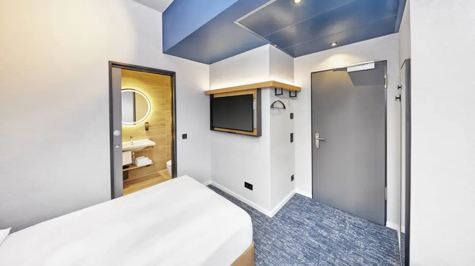 Einzelbett mit einem Fernseher gegenüber. Neben dem Bett ist das Badezimmer mit einem Waschbecken.