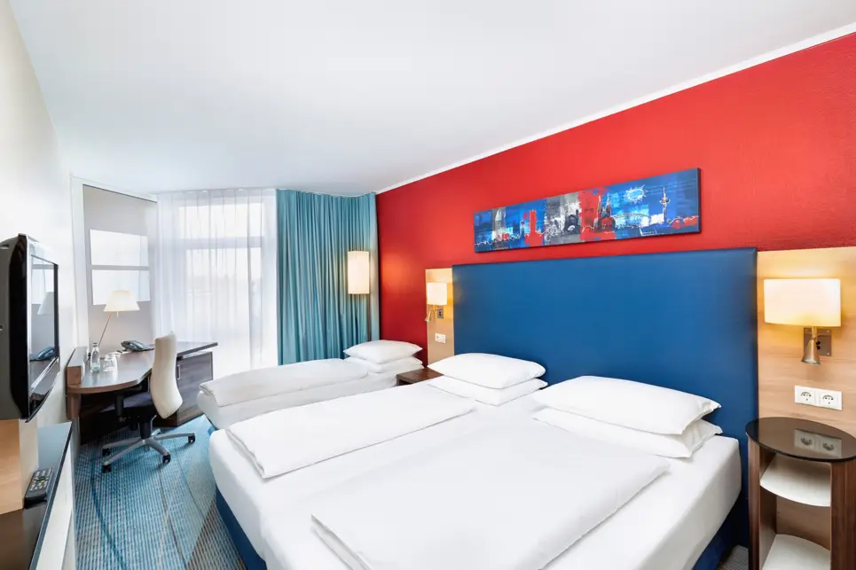 Doppelbett und ein Zustellbett im Hotelzimmer. Vor dem Fenster steht ein Schreibtisch.