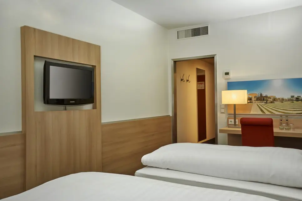 Twinbetten vor einem Fernseher an der Wand. Neben dem Bett steht ein Schreibtisch.