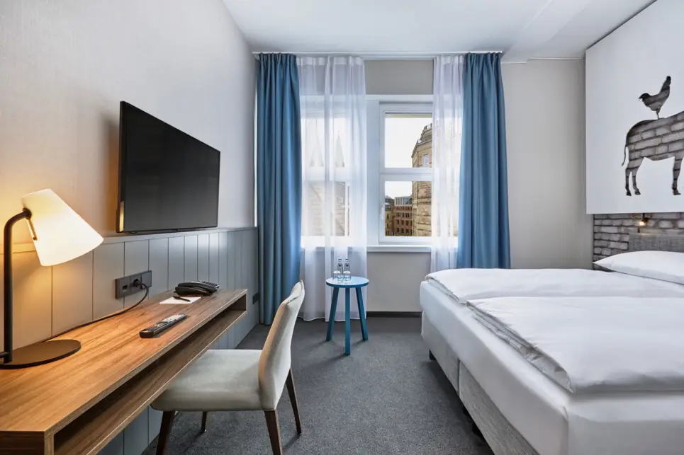 Hotelzimmer mit einem Doppelbett auf der rechten Seite und einem Schreibtisch auf der linken Seite. Über dem Schreibtisch hängt ein Fernseher.