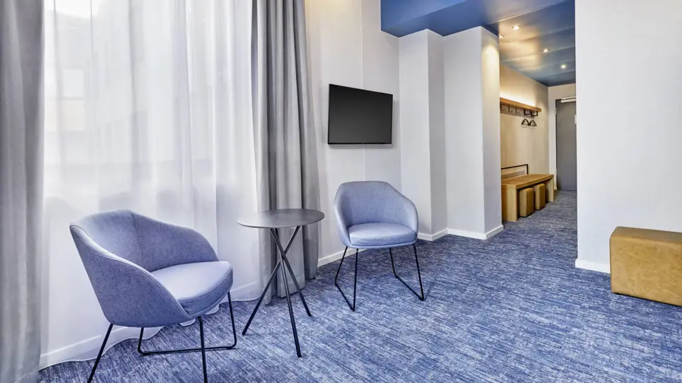 Zwei Stühle und ein Tisch stehen vor einem Fenster. In dem Hotelzimmerflur steht eine Ablagebank.