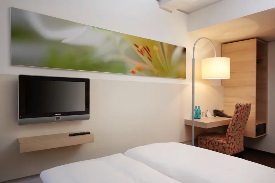 Blick vom Bett auf einen Fernseher und einem kleinen Schreibtisch. Neben dem Schreibtisch steht eine große Lampe.