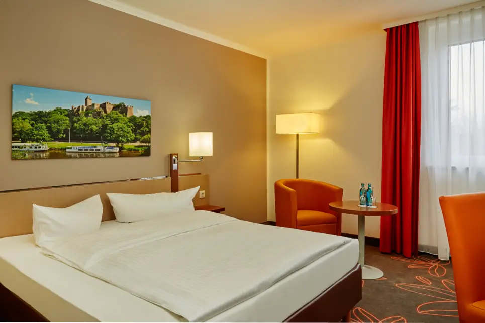 Hotelzimmer mit einem Doppelbett und einem Sessel. Über dem Bett hängt ein Bild.