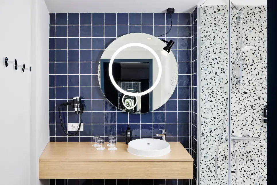 Waschtisch und eine Dusche im Badezimmer. Über dem Waschbecken hängt ein großer, runder Spiegel.