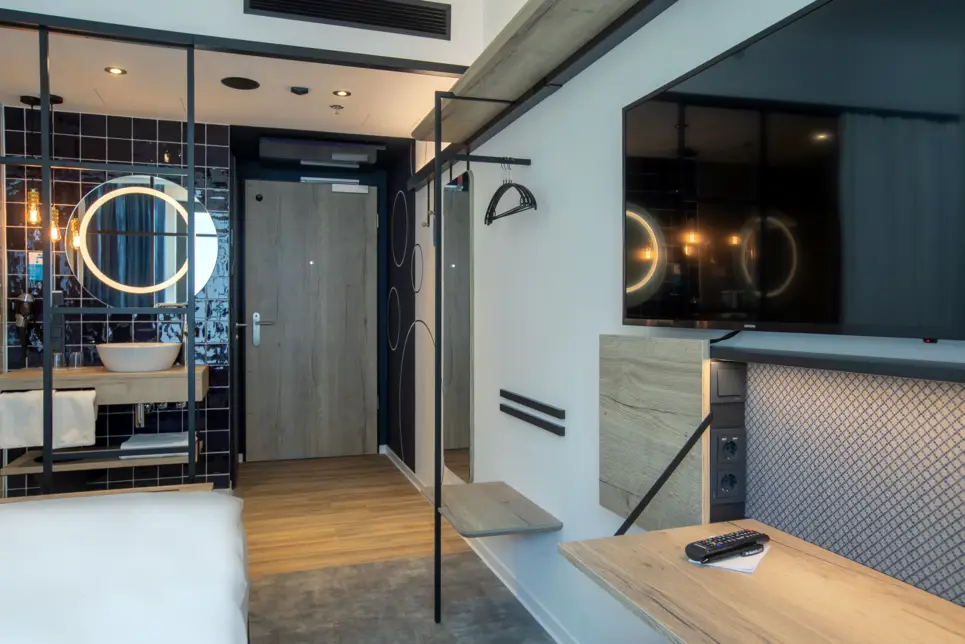 Im Vordergrund ist der Schreibtisch und ein Fernseher im Hotelzimmer. Im Hintergrund ist das offene Badezimmer mit einem Waschbecken.