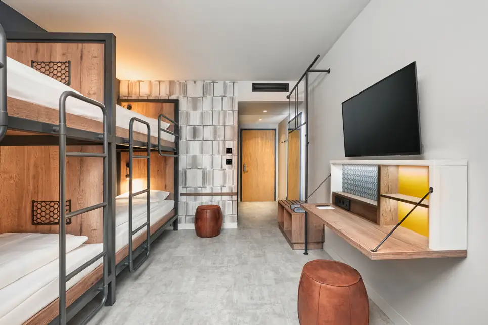 Hotelzimmer mit Etagenbetten, Hocker und einer Ablagebank. Gegenüber der Betten hängt ein Fernseher und ein Schreibtisch.