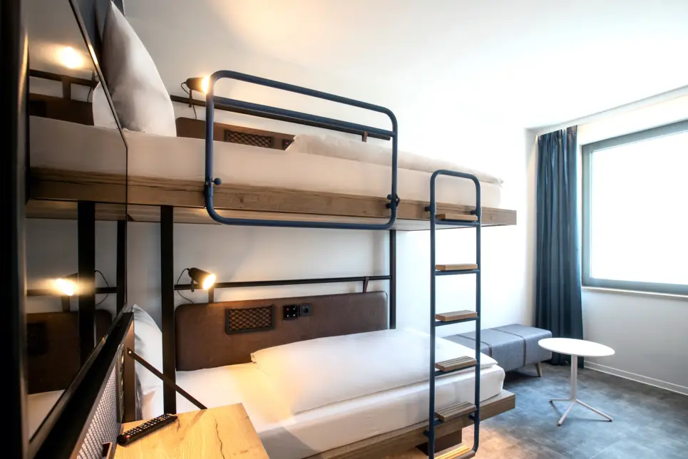 Helles Hotelzimmer mit Etagenbetten. Vor dem Fenster ist eine kleine Sitzgruppe.