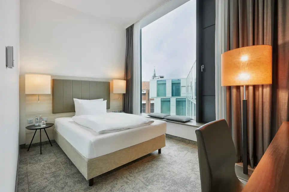 Einzelbett vor einem großen Fenster mit einer kleinen Sitzbank. Auf der rechten Seite steht eine große Stehlampe.