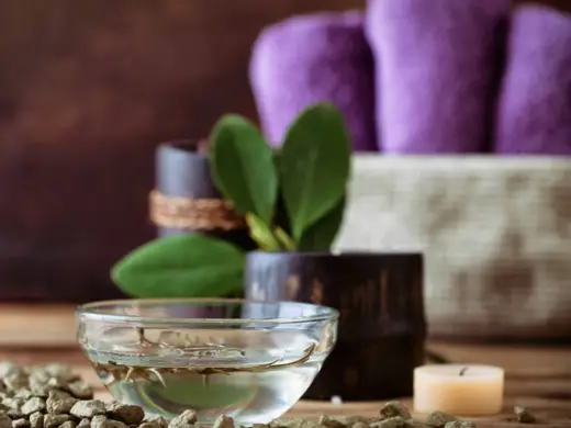 Nahaufnahme von einem Öl in einer Schale. Im Hintergrund sind verschwommen ein Bündel lila Handtüscher