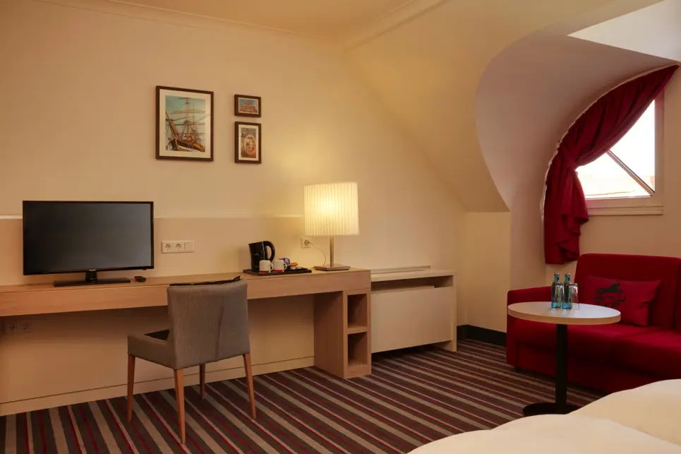 Schreibtisch mit einem Stuhl und einem Fernseher im Hotelzimmer. Auf der rechten Seite ist eine Sitzecke.