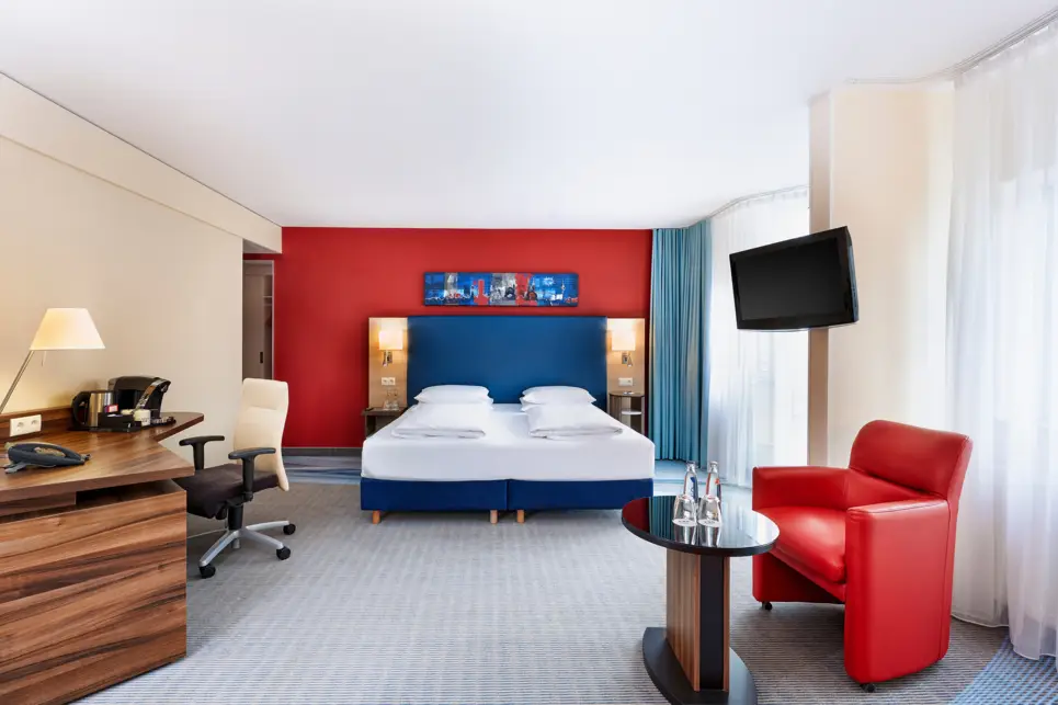 Großes Hotelzimmer mit einer Sitzecke rechts und einem Schreibtisch links. In der Mitte steht ein Doppelbett.