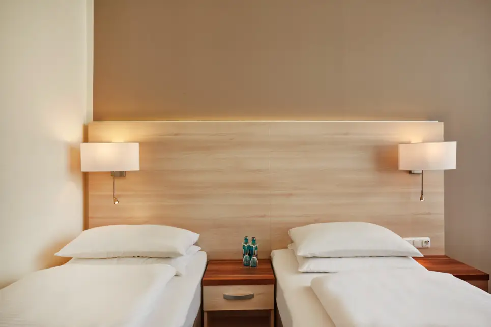 Zwei Einzelbetten mit Nachttisch zwischen den Betten. Auf dem Nachttisch stehen 2 Wasserflaschen und 2 Gläser.