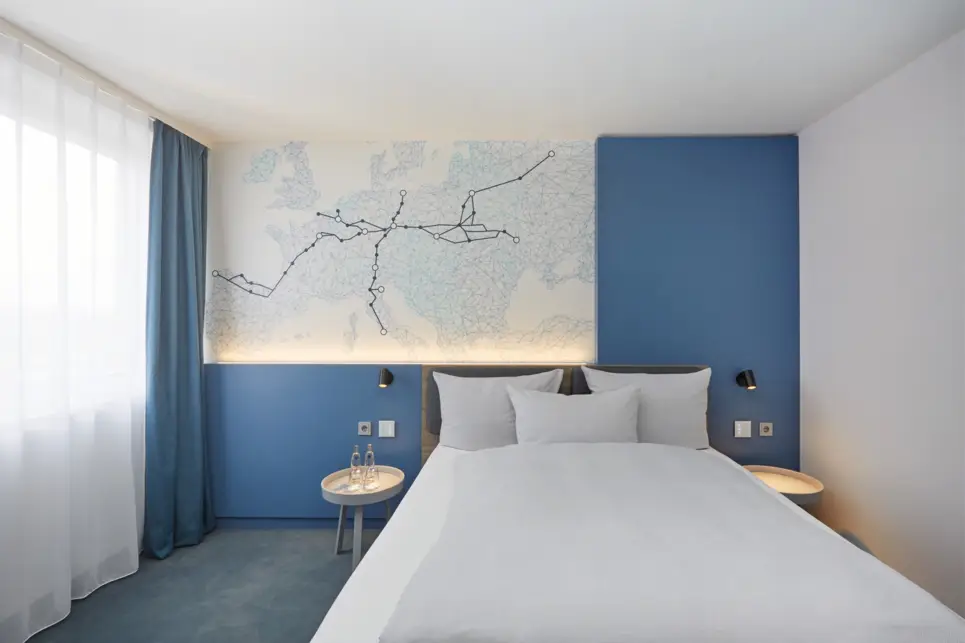 Einzelbett an einer blauen Wand mit einem großen Wandbild. Das Bild zeigt die Europakarte auf der mehrere schwarze Linien von Leipzig weggehen.