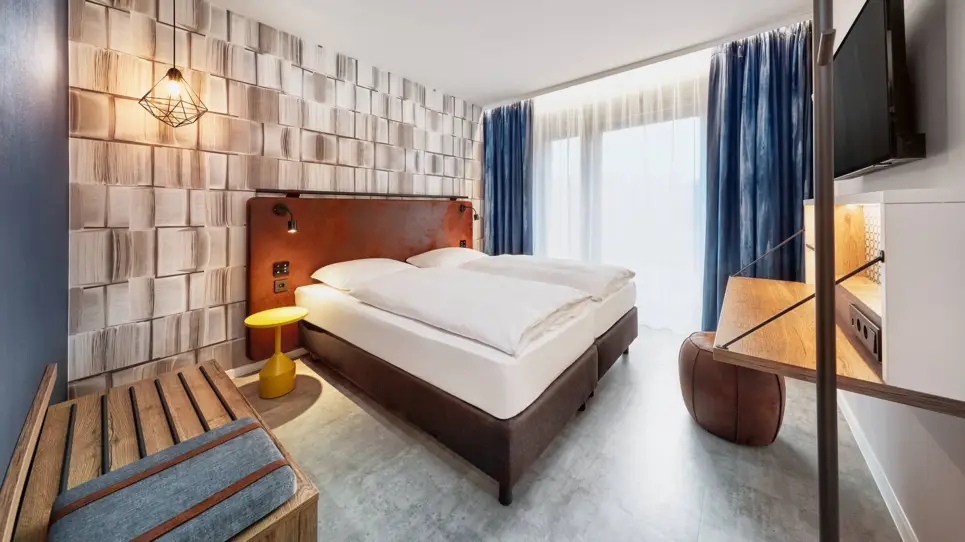 Doppelbett in einem Hotelzimmer mit einer Ablagebank auf einer Seite und einem Schreibtisch auf der anderen Seite.