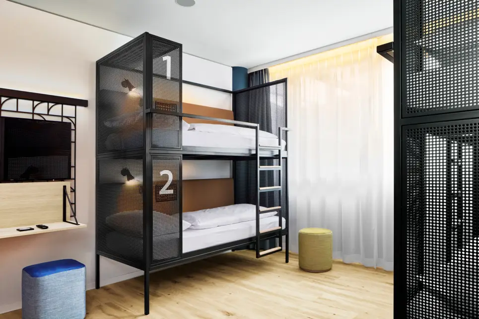 Etagenbett in einem Hotelzimmer vor einem Fenster. Neben dem Bett steht ein Hocker, ein Schreibtisch und ein Fernseher.