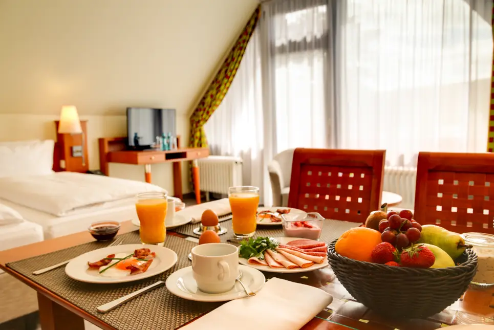 Reichlich gedeckter Frühstückszisch auf dem Hotelzimmer. Im Hintergrund steht ein Doppelbett.