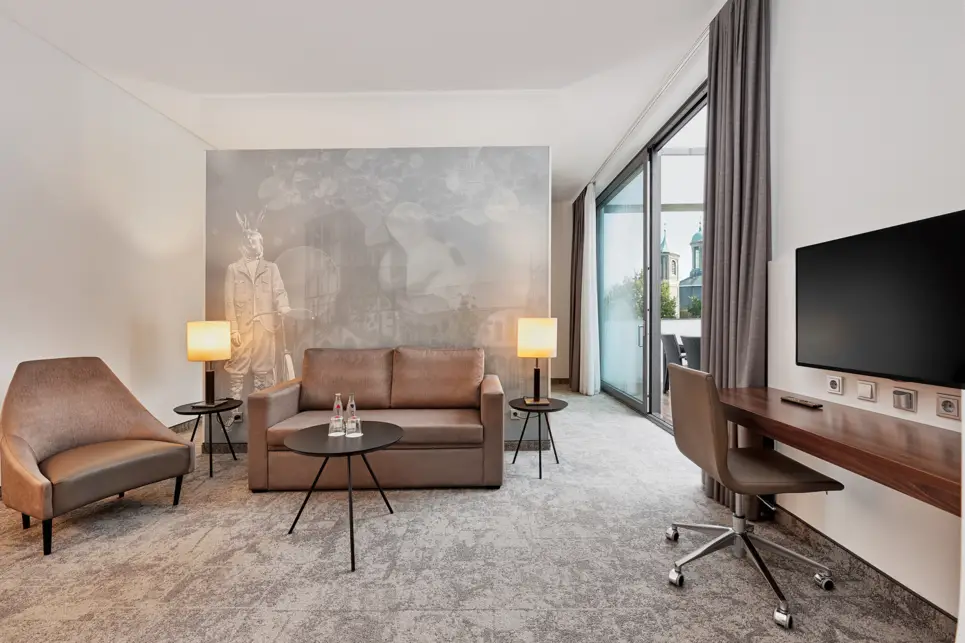 Wohnbereich mit einer braunen Couch und einem braunen Sessel. Auf der rechten Seite ist ein Schreibtischm it einem Fernseher.